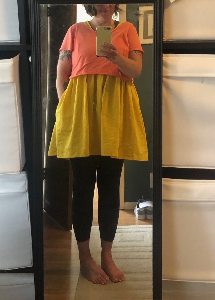 A mirror selfie of me, in the yellow dress with a salmony pink tee shirt over it and black leggings underneath. I have a smirk on my face, because of course I do