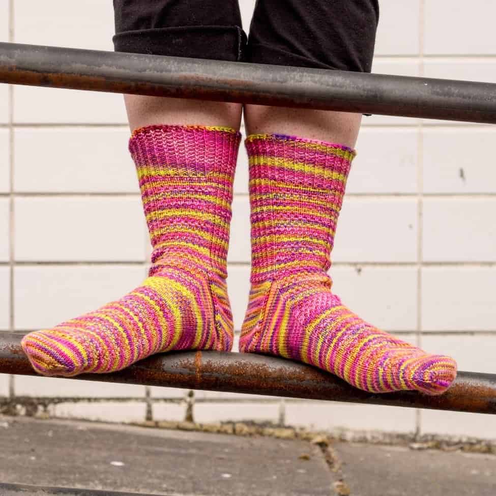 two pink-hand-knit-clad feet stand on a railing, toes pointed outward